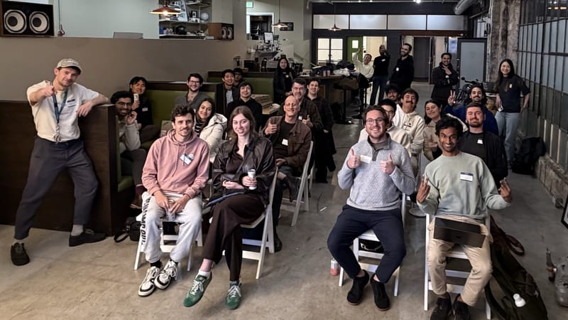 Group of about 25 people smiling and giving thumbs up in an industrial loft venue at a Claude Code Show and Tell meetup