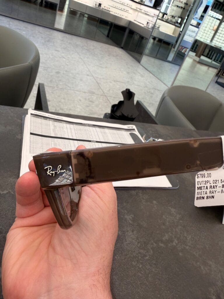 Hand holding a brown Meta Ray-Ban display temple piece showing the Ray-Ban logo, with a price tag reading $799 in an eyewear retail store