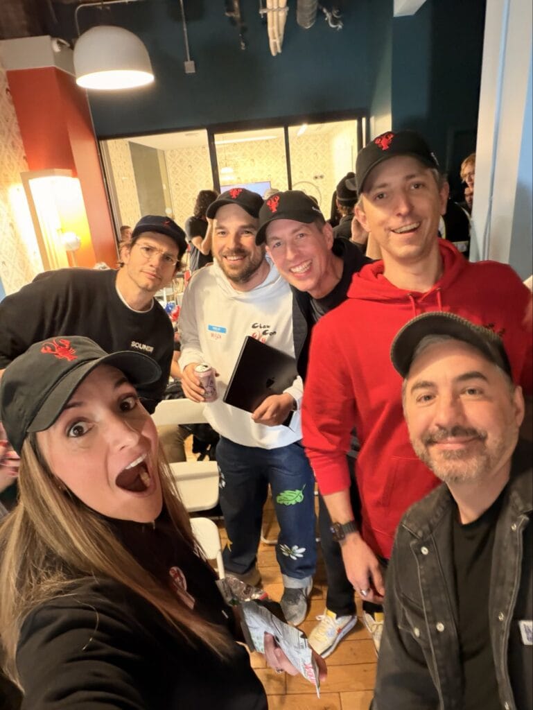 Group selfie of six ClawCon attendees wearing black hats with a red lobster logo at a crowded indoor venue, with one person in a white ClawCon hoodie holding a tablet