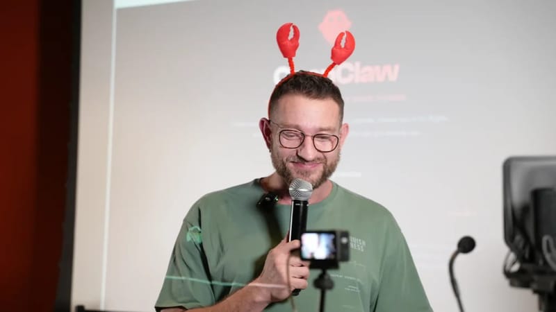 Man wearing lobster claw headband and glasses speaking into a microphone at ClawCon, with the ClawCon logo visible on a screen behind him