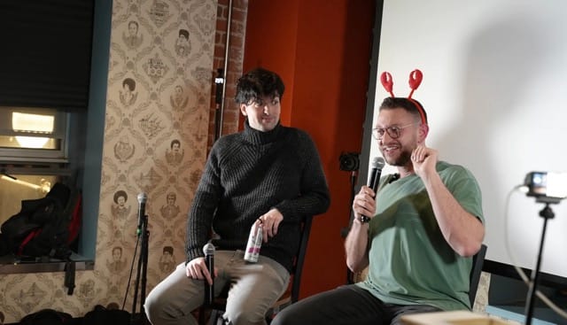 Two men seated on chairs at a ClawCon panel discussion; the man on the right wears lobster claw headbands and holds a microphone while the man on the left sits in a black turtleneck